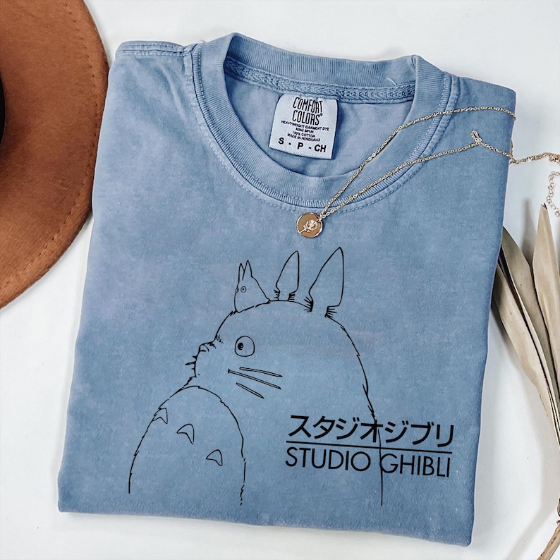 May include: A light blue t-shirt featuring a black graphic of a cartoon character and Japanese text. The text reads "STUDIO GHIBLI". A gold necklace with a pendant is draped across the shirt. The shirt is labeled "COMFORT COLORS".