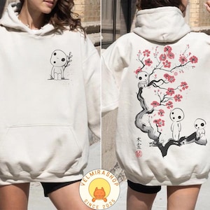 May include: Cream-colored hoodie featuring a front pocket and a drawstring hood. The front displays a small black and white illustration of a forest spirit holding a sprig. The back showcases a larger design of a tree with pink blossoms and more forest spirits.