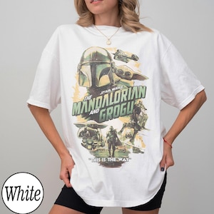 May include: White t-shirt featuring a graphic from "The Mandalorian" with the text "The Mandalorian and Grogu" and the phrase "This is the Way." The design includes images of the Mandalorian, Grogu, and starships.