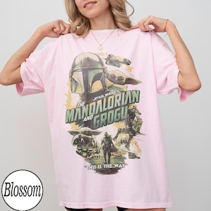 May include: A light pink t-shirt featuring a Star Wars graphic with The Mandalorian and Grogu. The design includes the Mandalorian's helmet, Grogu, and the phrase "This is the Way" in green and white.