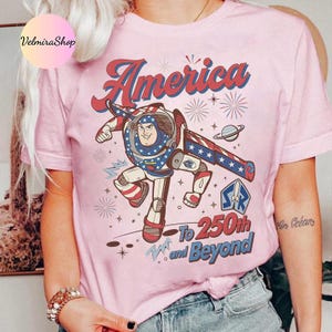 May include: Pink t-shirt featuring a cartoon illustration of a space ranger with patriotic colors and the word "America" in red script. The shirt also includes the text "To 250th and Beyond."