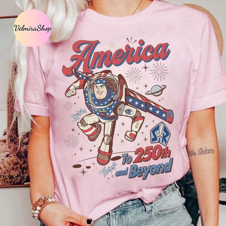 May include: Pink t-shirt featuring a cartoon illustration of a space ranger with patriotic colors and the word "America" in red script. The shirt also includes the text "To 250th and Beyond."