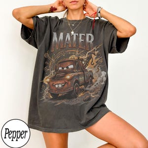 May include: Dark gray t-shirt featuring a graphic of Mater from the movie Cars. The word "MATER" is printed above the character. The design includes a brown tow truck with a smiling face, set against a background of gears and lightning.