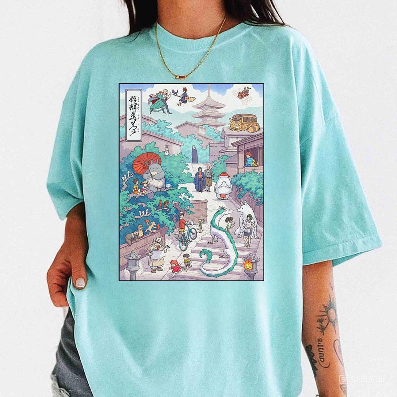 May include: A light blue t-shirt featuring a colorful illustration of a Japanese street scene with animated characters. The graphic includes a pagoda, a dragon, and various figures in a detailed, whimsical design. The shirt has a relaxed fit.