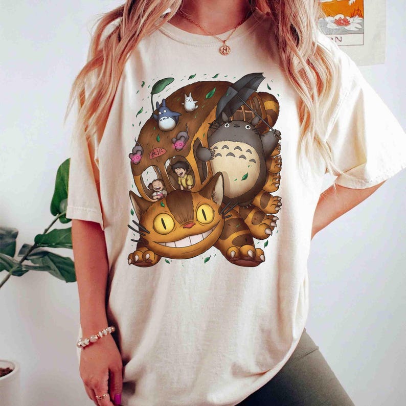 May include: Cream-colored t-shirt featuring a colorful graphic of characters from the animated film "My Neighbor Totoro." The design includes Totoro, a cat bus, and other characters. The shirt has a crew neck and short sleeves.