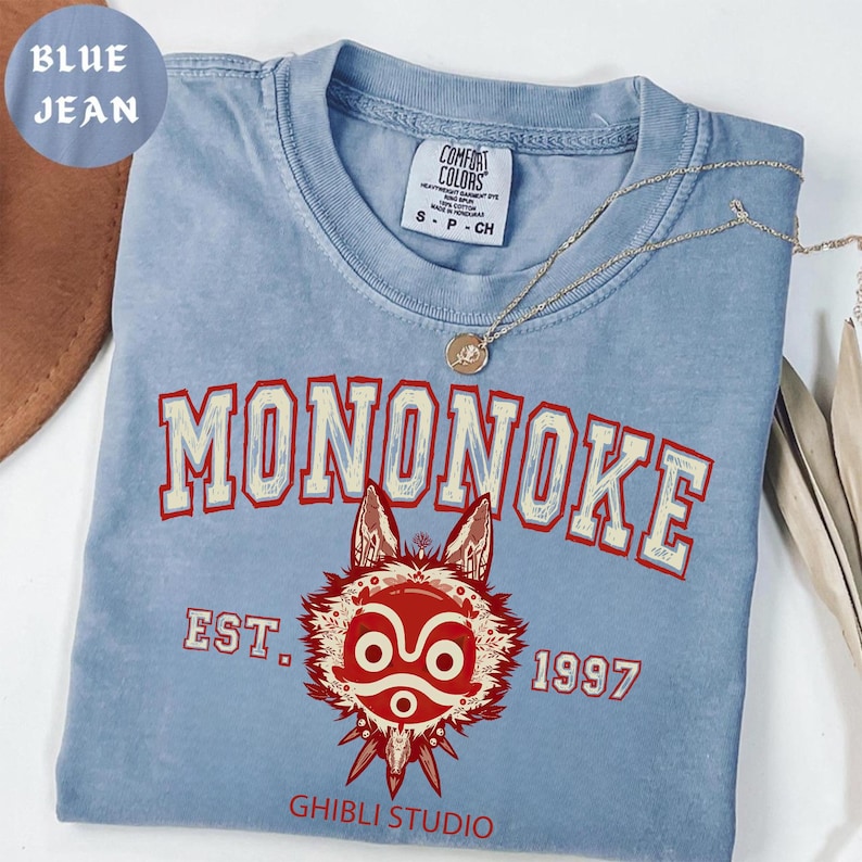 May include: A light blue Comfort Colors t-shirt with the word "MONONOKE" in red and white lettering. Below the word is a red and white graphic of a wolf-like creature. The text "EST. 1997" and "GHIBLI STUDIO" are also visible.