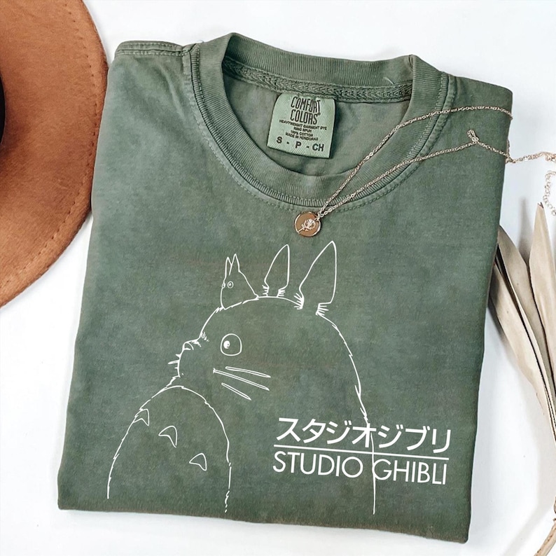May include: Olive green t-shirt featuring a white graphic of a cartoon character and the text "STUDIO GHIBLI" in English and Japanese. A gold-colored necklace with a pendant rests on the shirt. The shirt is folded and has a tag that says "COMFORT COLORS".