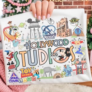 Può includere: T-shirt bianca con illustrazioni colorate delle attrazioni e dei personaggi di Disney's Hollywood Studios. Include Topolino, i personaggi di Toy Story e l'Hollywood Tower Hotel. Le parole "Hollywood Studios" sono ben visibili.