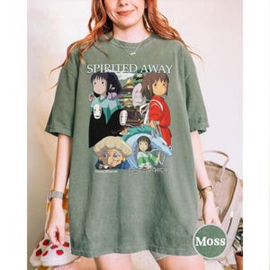 Pode incluir: T-shirt verde musgo com um gr&aacute;fico do filme de anima&ccedil;&atilde;o "A Viagem de Chihiro". O design inclui personagens e o t&iacute;tulo do filme. O texto "Hayao Miyazaki" e texto japon&ecirc;s tamb&eacute;m s&atilde;o vis&iacute;veis. A camisa tem um ajuste relaxado.