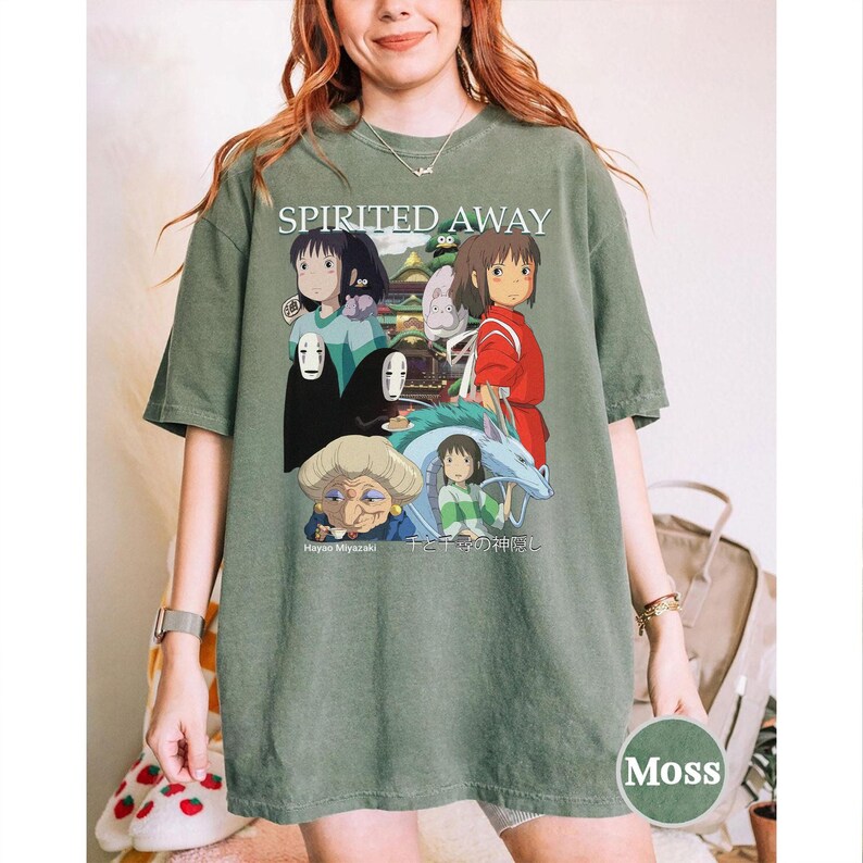 Pode incluir: T-shirt verde musgo com um gr&aacute;fico do filme de anima&ccedil;&atilde;o "A Viagem de Chihiro". O design inclui personagens e o t&iacute;tulo do filme. O texto "Hayao Miyazaki" e texto japon&ecirc;s tamb&eacute;m s&atilde;o vis&iacute;veis. A camisa tem um ajuste relaxado.