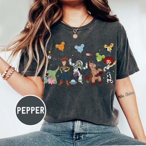 Puede incluir: Camiseta gris oscuro con un gr&aacute;fico colorido de personajes de Toy Story, incluyendo a Woody y Buzz Lightyear. El dise&ntilde;o incluye im&aacute;genes con forma de globo y la palabra "Pepper" en un c&iacute;rculo.