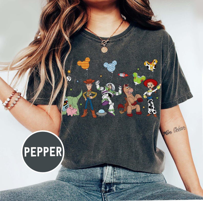 Puede incluir: Camiseta gris oscuro con un gr&aacute;fico colorido de personajes de Toy Story, incluyendo a Woody y Buzz Lightyear. El dise&ntilde;o incluye im&aacute;genes con forma de globo y la palabra "Pepper" en un c&iacute;rculo.