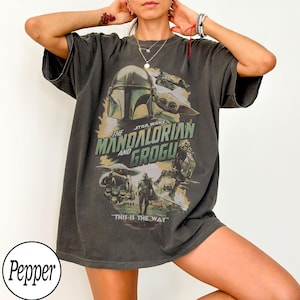 May include: Dark gray t-shirt featuring a graphic from the Star Wars series, "The Mandalorian and Grogu." The design includes images of the Mandalorian, Grogu, and the text "THIS IS THE WAY."