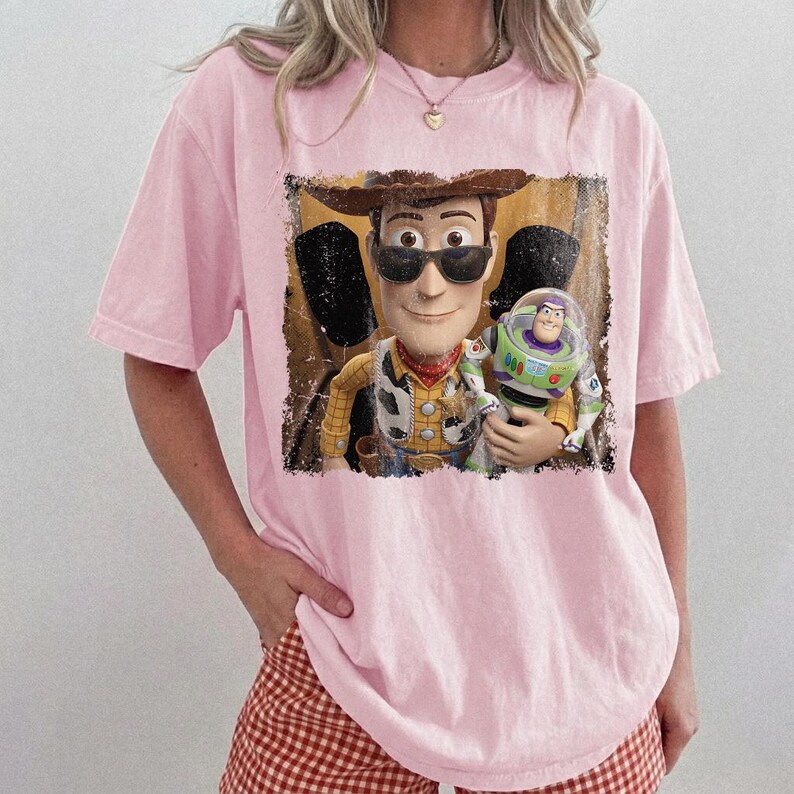 Op de afbeelding: Een lichtroze T-shirt met korte mouwen en een grafische print van een cowboy-personage met een zwarte zonnebril die een space ranger-actiefiguur vasthoudt. De afbeelding heeft een verweerd, getextureerd uiterlijk met een donkere rand.