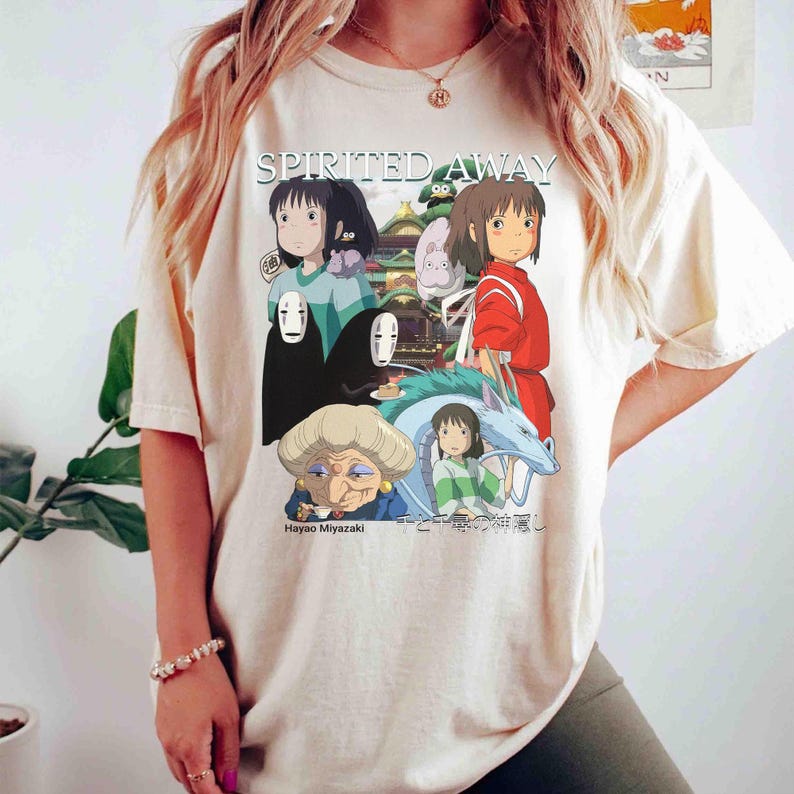 Pode incluir: Camiseta creme com um gr&aacute;fico do filme de anima&ccedil;&atilde;o "A Viagem de Chihiro". O design inclui personagens e o t&iacute;tulo do filme em branco. A parte inferior do gr&aacute;fico inclui o texto "Hayao Miyazaki" e texto japon&ecirc;s.