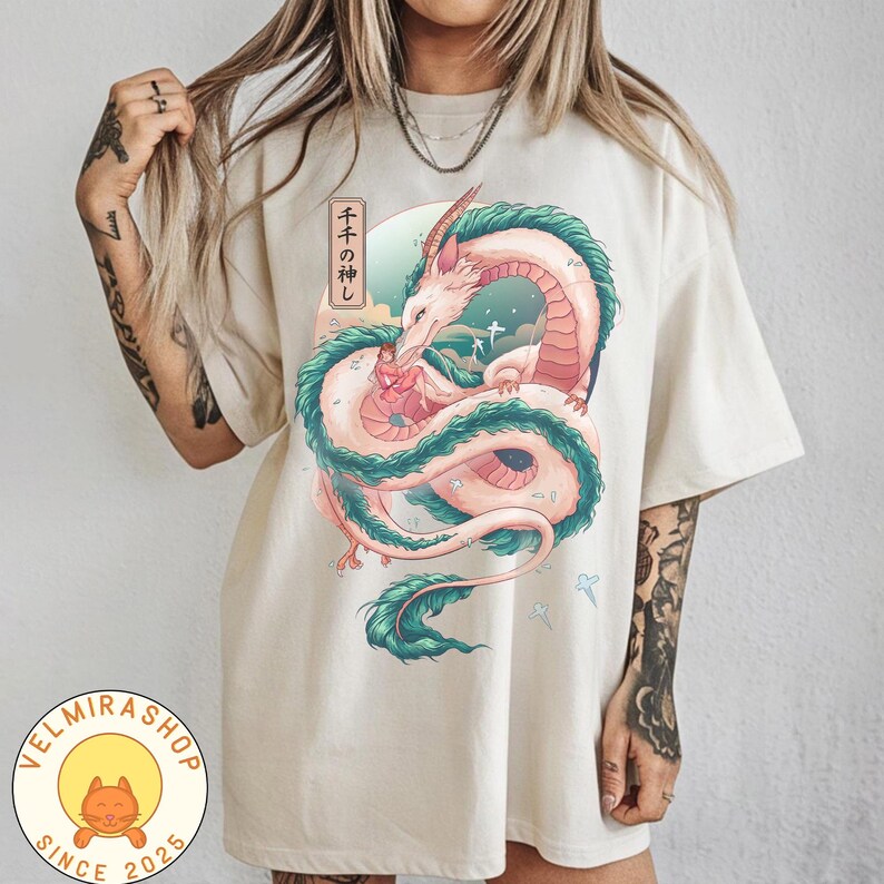 May include: Cream-colored t-shirt featuring a colorful illustration of a dragon with the text "千と千尋の神隠し" in Japanese. The dragon is depicted in shades of pink, green, and white. The shirt also has the shop's logo in the bottom left corner.