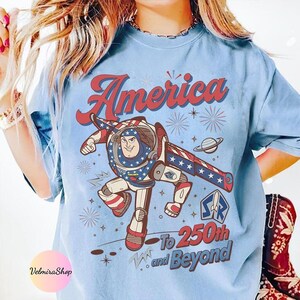 May include: Light blue t-shirt featuring a graphic of a cartoon astronaut with the text "America" and "To 250th and Beyond". The astronaut is in a patriotic design with red, white, and blue colors. The shirt has a vintage, distressed look.