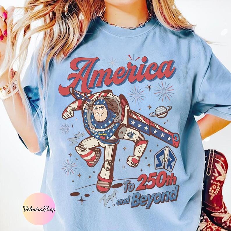May include: Light blue t-shirt featuring a graphic of a cartoon astronaut with the text "America" and "To 250th and Beyond". The astronaut is in a patriotic design with red, white, and blue colors. The shirt has a vintage, distressed look.