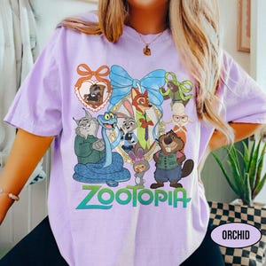 Puede incluir: Camiseta de color orqu&iacute;dea con un gr&aacute;fico colorido de personajes de la pel&iacute;cula "Zootopia". El dise&ntilde;o incluye un lazo azul, un marco en forma de coraz&oacute;n y la palabra "Zootopia" en verde y azul. La camiseta tiene mangas cortas.