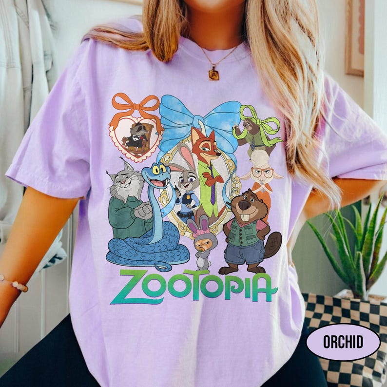 Puede incluir: Camiseta de color orqu&iacute;dea con un gr&aacute;fico colorido de personajes de la pel&iacute;cula "Zootopia". El dise&ntilde;o incluye un lazo azul, un marco en forma de coraz&oacute;n y la palabra "Zootopia" en verde y azul. La camiseta tiene mangas cortas.
