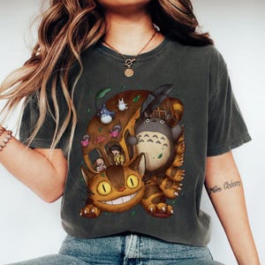 May include: Dark gray t-shirt featuring a colorful graphic of characters from the animated film "My Neighbor Totoro." The design includes Totoro, the Catbus, and other forest spirits. The shirt has a relaxed fit and short sleeves.