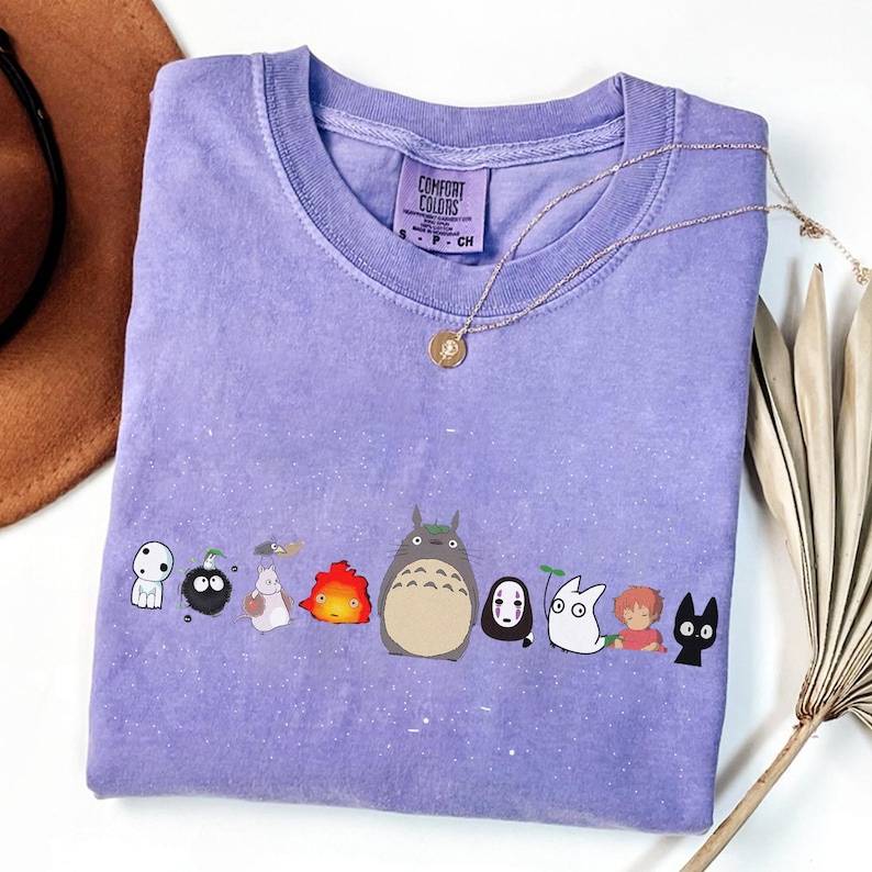 May include: A lavender Comfort Colors t-shirt featuring a line of cartoon characters. The characters include a large gray creature, a black cat, and other colorful figures. A gold necklace with a pendant is draped across the shirt.