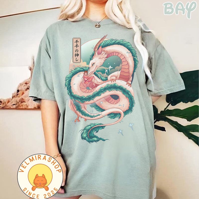 May include: Sage green t-shirt featuring a detailed illustration of a dragon with pink, white, and teal accents. The dragon is intertwined with a Japanese text box. The shirt has a relaxed fit and short sleeves.