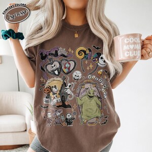 Puede incluir: Una camiseta marr&oacute;n de manga corta con un estampado gr&aacute;fico de personajes y s&iacute;mbolos de Pesadilla antes de Navidad, incluyendo a Jack Skellington, Sally, Oogie Boogie y Zero, con un lazo morado y detalles de telara&ntilde;as.