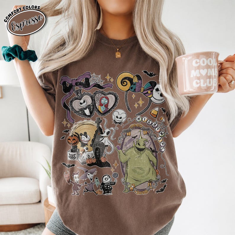Puede incluir: Una camiseta marr&oacute;n de manga corta con un estampado gr&aacute;fico de personajes y s&iacute;mbolos de Pesadilla antes de Navidad, incluyendo a Jack Skellington, Sally, Oogie Boogie y Zero, con un lazo morado y detalles de telara&ntilde;as.