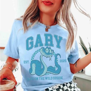 May include: Light blue t-shirt with the name "GARY" in large, block letters. Below the name is a cartoon snake illustration. The text "EST. 2025" and "LIFE ON THE WILD SSSIDE" are also printed on the shirt.