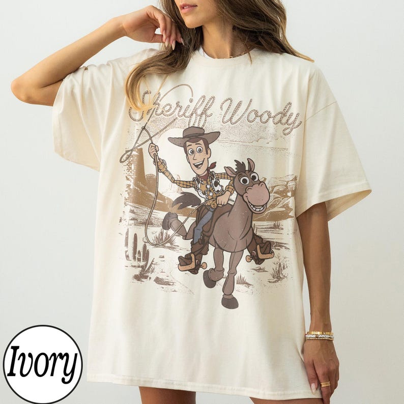 Op de afbeelding: Ivoor kleurig T-shirt met een afbeelding van Sheriff Woody op een paard, met de tekst "Sheriff Woody" erboven. Het ontwerp gebruikt bruine en beige tinten, tegen een woestijnlandschap. Het T-shirt heeft korte mouwen.
