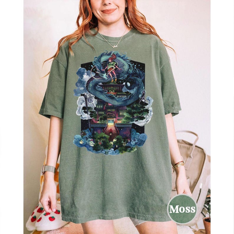 Puede incluir: Camiseta verde musgo con un gr&aacute;fico colorido de un drag&oacute;n y una persona en un edificio, rodeados de nubes y agua. La camiseta tiene mangas cortas y cuello redondo. La palabra "Moss" est&aacute; impresa en una etiqueta circular.