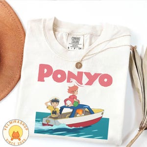 May include: Cream-coloured t-shirt with the pink word "PONYO" and a cartoon illustration of characters in a boat. The graphic shows two children in a boat on the water. A gold necklace and a brown hat are also visible.