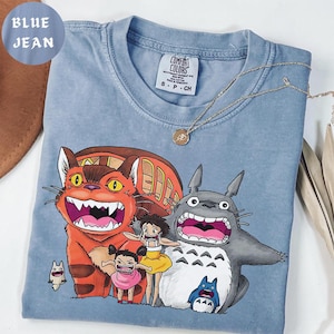 May include: Blue jean colored t-shirt featuring a graphic of characters from the animated film, My Neighbor Totoro. The shirt has a crew neck and short sleeves. A gold necklace is draped across the shirt.