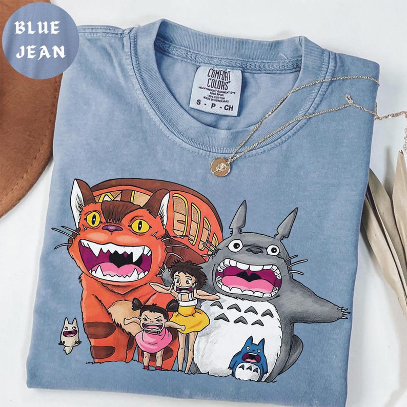 May include: Blue jean colored t-shirt featuring a graphic of characters from the animated film, My Neighbor Totoro. The shirt has a crew neck and short sleeves. A gold necklace is draped across the shirt.