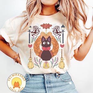 May include: Cream-colored t-shirt featuring a cartoon black cat with a red bow tie, surrounded by a braided wreath, flowers, and witch-themed elements. The shirt has a round neck and short sleeves. The shop logo is in the bottom left corner.