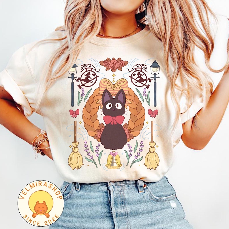 May include: Cream-colored t-shirt featuring a cartoon black cat with a red bow tie, surrounded by a braided wreath, flowers, and witch-themed elements. The shirt has a round neck and short sleeves. The shop logo is in the bottom left corner.