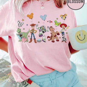 Puede incluir: Una camiseta rosa claro con un dise&ntilde;o colorido de personajes de Toy Story y globos. La camiseta incluye im&aacute;genes de Woody, Buzz Lightyear y otros personajes. La palabra "Blossom" est&aacute; impresa en la esquina superior derecha.
