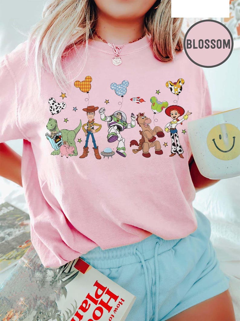 Puede incluir: Una camiseta rosa claro con un dise&ntilde;o colorido de personajes de Toy Story y globos. La camiseta incluye im&aacute;genes de Woody, Buzz Lightyear y otros personajes. La palabra "Blossom" est&aacute; impresa en la esquina superior derecha.