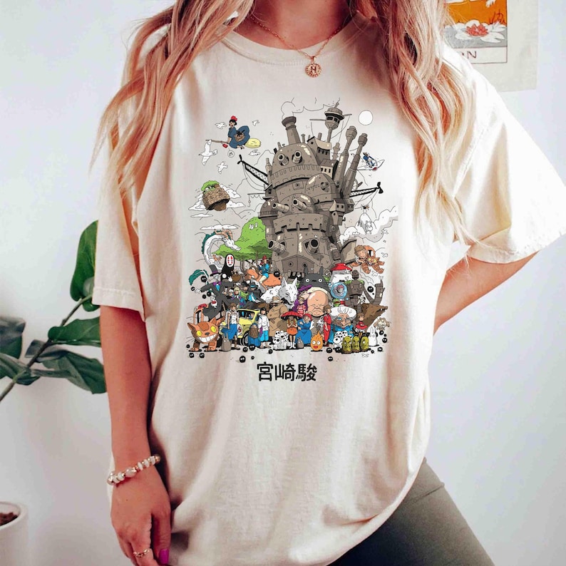 以下が含まれることがあります： クリーム色のTシャツ。城と様々な漫画のキャラクターの詳細なイラストが描かれています。グラフィックには、下部に日本語のテキストが含まれています。シャツは柔らかい素材で作られています。