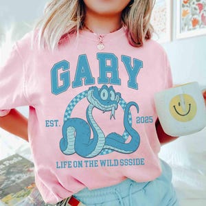 May include: A light pink t-shirt with the name "GARY" in blue block letters above a cartoon blue snake. The snake is inside a checkered circle, with "EST. 2025" and "LIFE ON THE WILD SSSIDE" printed on the shirt.