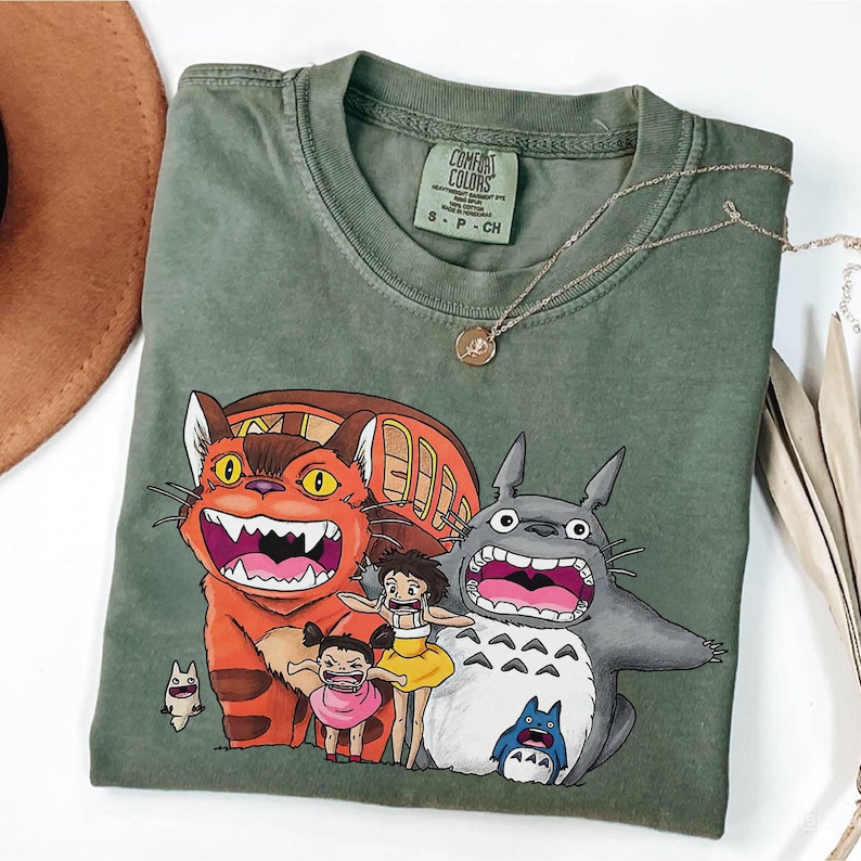 May include: Olive green Comfort Colors t-shirt featuring a colorful graphic of characters from the animated film, My Neighbor Totoro. The design includes a large orange cat, Totoro, and two children. A gold necklace is draped across the shirt.