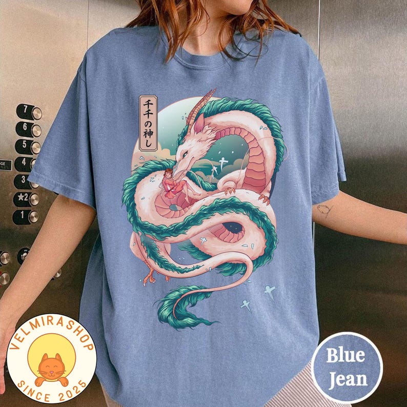 May include: Blue jean colored t-shirt featuring a dragon illustration with Japanese text. The dragon is white and pink with teal accents, set against a backdrop of a blue sky and clouds. The shirt has a round neck and short sleeves.