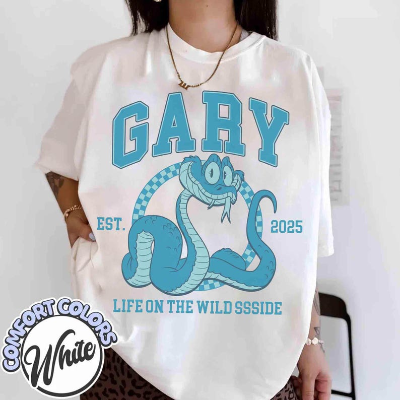 May include: White Comfort Colors t-shirt with a blue snake graphic. The word "GARY" is arched above the snake, with "EST. 2025" and "LIFE ON THE WILD SSSSIDE" below. The snake is inside a checkered circle.