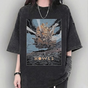 Puede incluir: Camiseta gris oscuro con un gr&aacute;fico de "El castillo ambulante". El dise&ntilde;o muestra el castillo en tonos marrones, naranjas y azules, sobre un cielo nublado. El texto "HOWL'S MOVING CASTLE" est&aacute; impreso debajo.