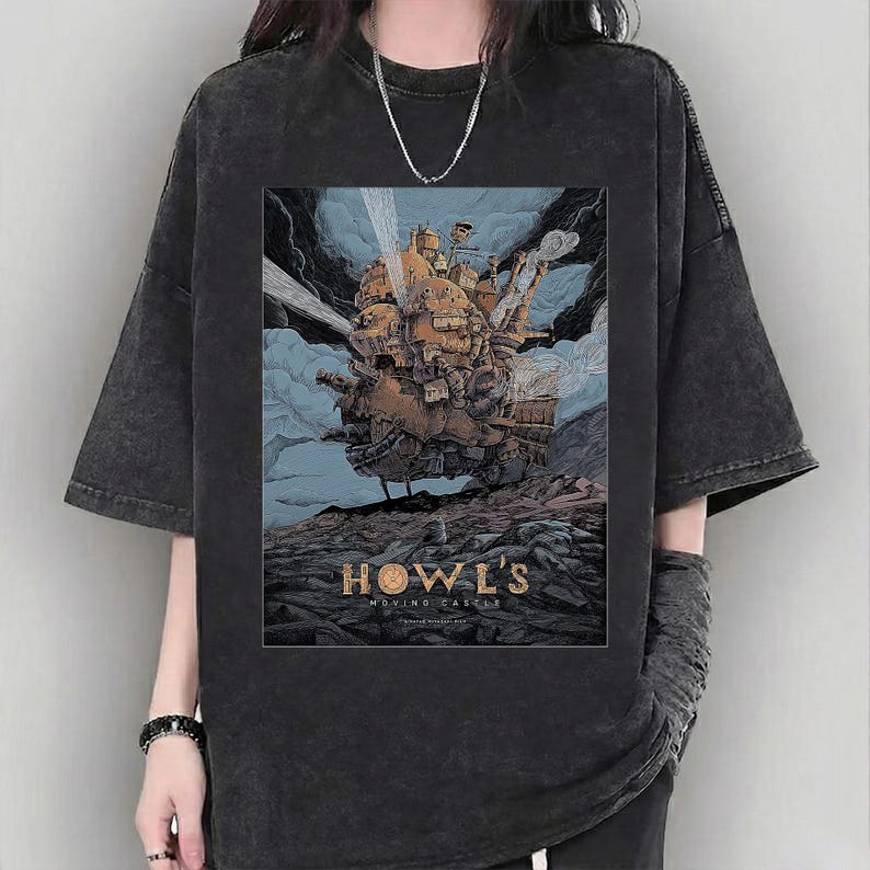 Puede incluir: Camiseta gris oscuro con un gr&aacute;fico de "El castillo ambulante". El dise&ntilde;o muestra el castillo en tonos marrones, naranjas y azules, sobre un cielo nublado. El texto "HOWL'S MOVING CASTLE" est&aacute; impreso debajo.