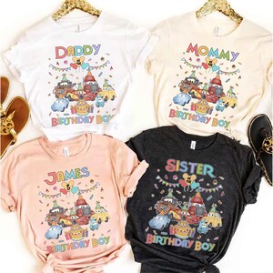 Puede incluir: Cuatro camisetas de manga corta con gr&aacute;ficos coloridos. Las camisetas son blancas, beige, rosas y gris oscuro. Cada camiseta presenta un dise&ntilde;o de coche de dibujos animados y las palabras "Daddy", "Mommy", "James" y "Sister" con "Birthday Boy" debajo.