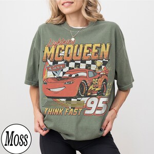 K&ouml;nnte beinhalten: Olivgr&uuml;nes T-Shirt mit einer Lightning McQueen-Grafik aus dem Film Cars. Das Design enth&auml;lt den Text "Lightning McQueen", "Cars", "Think Fast" und die Nummer "95". Das T-Shirt hat einen Vintage-Look.