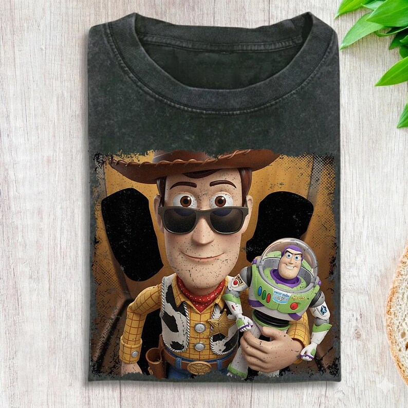 Op de afbeelding: Een antracietgrijs acid-wash T-shirt met een grafische print van het personage Woody met een zonnebril op, die een Buzz Lightyear-actiefiguur vasthoudt. Het ontwerp staat op een gele en zwarte achtergrond met een verweerde, textuurrijke uitstraling.
