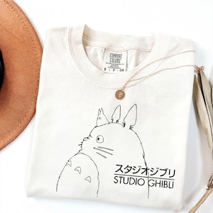 May include: Cream-colored t-shirt featuring a black line drawing of a cartoon character with the Japanese text "スタジオジブリ STUDIO GHIBLI". A gold necklace with a pendant is draped across the shirt. The shirt is folded, with a brown hat visible in the background.